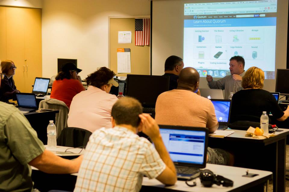 A man lectures from the front of a classroom full of people sitting in front of computers.