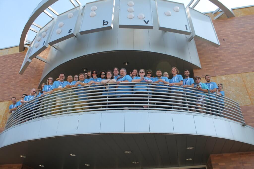 A group of 35 people stand look down, over the edge of a rounded balcony. Above them is a large sign reading "tsbvi."