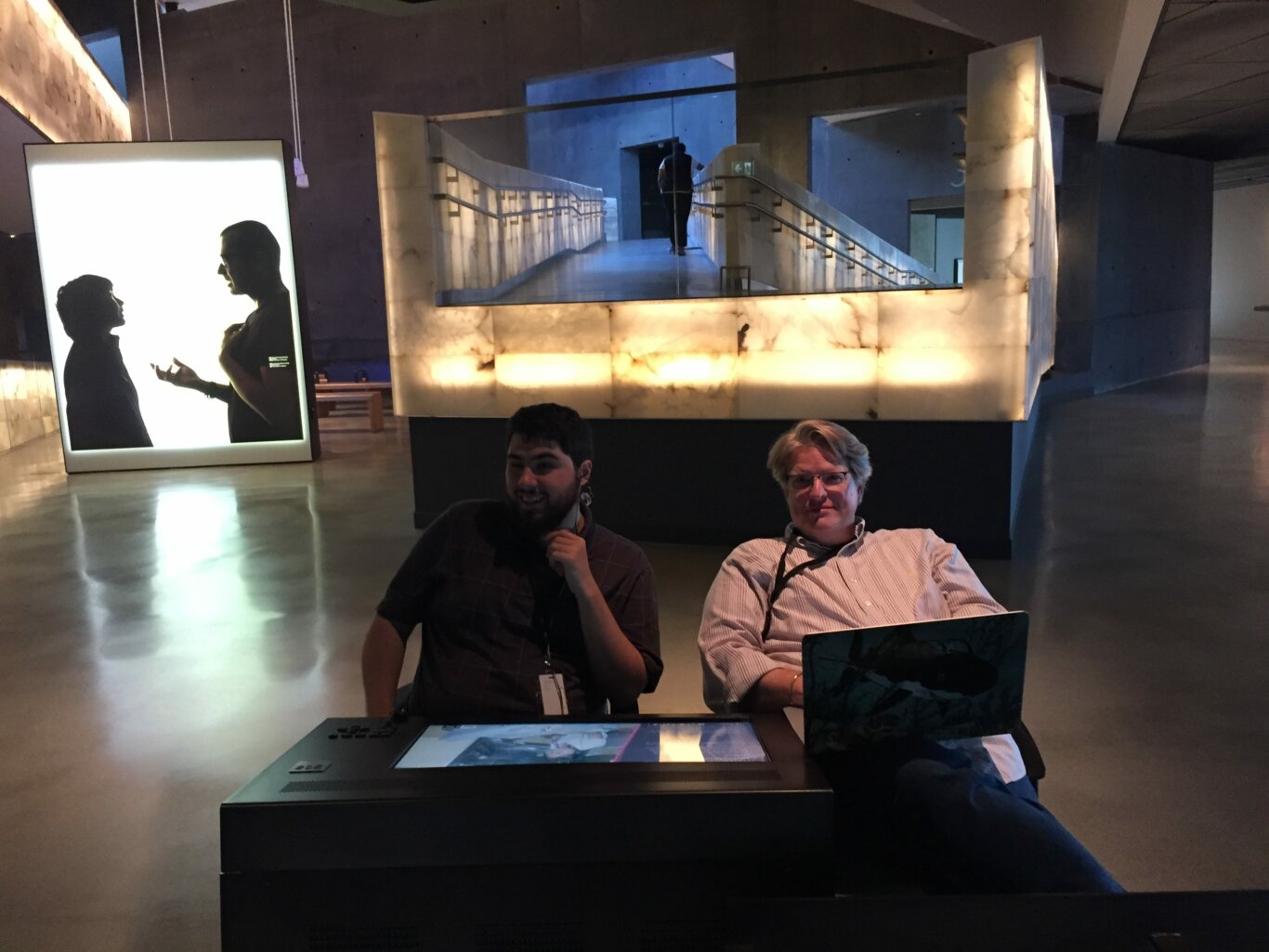 Two men sit at a table in a dimly lit space in front of a projection and a large, illuminated ramp.