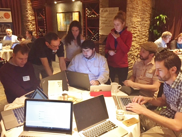 A small crowd hovers behind and around Sina coding at table, illuminated by his laptop screen.