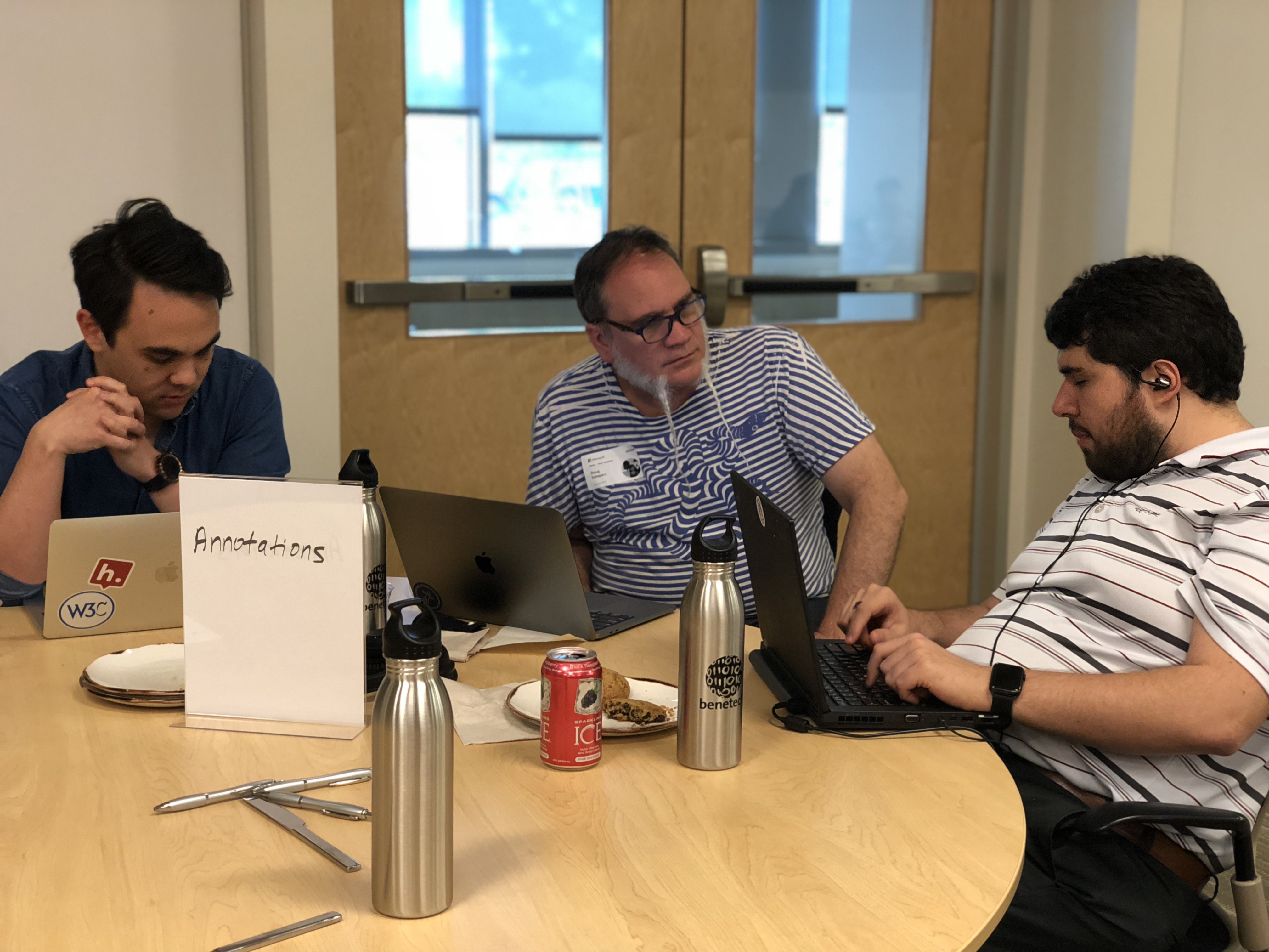 Sina sitting a table with two men; one man with a long beard formed into two points converses with Sina about an aspect of a project while the other man listens in.
