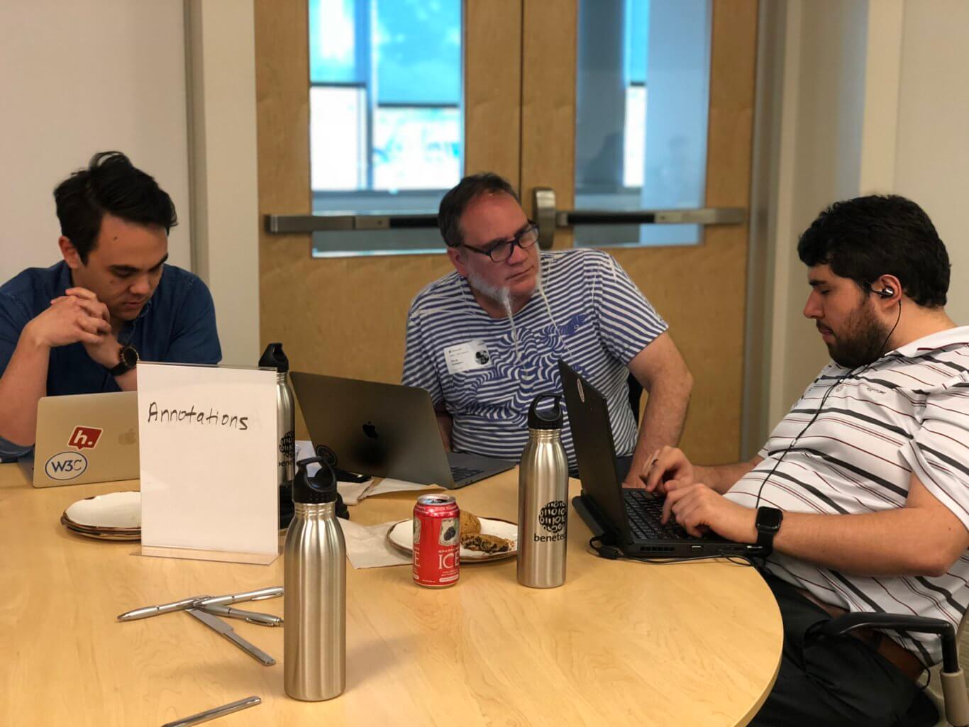 Sina sits a table with two men at a round table; one man with a long beard formed into two points is in conversation with Sina while the other man listens in.