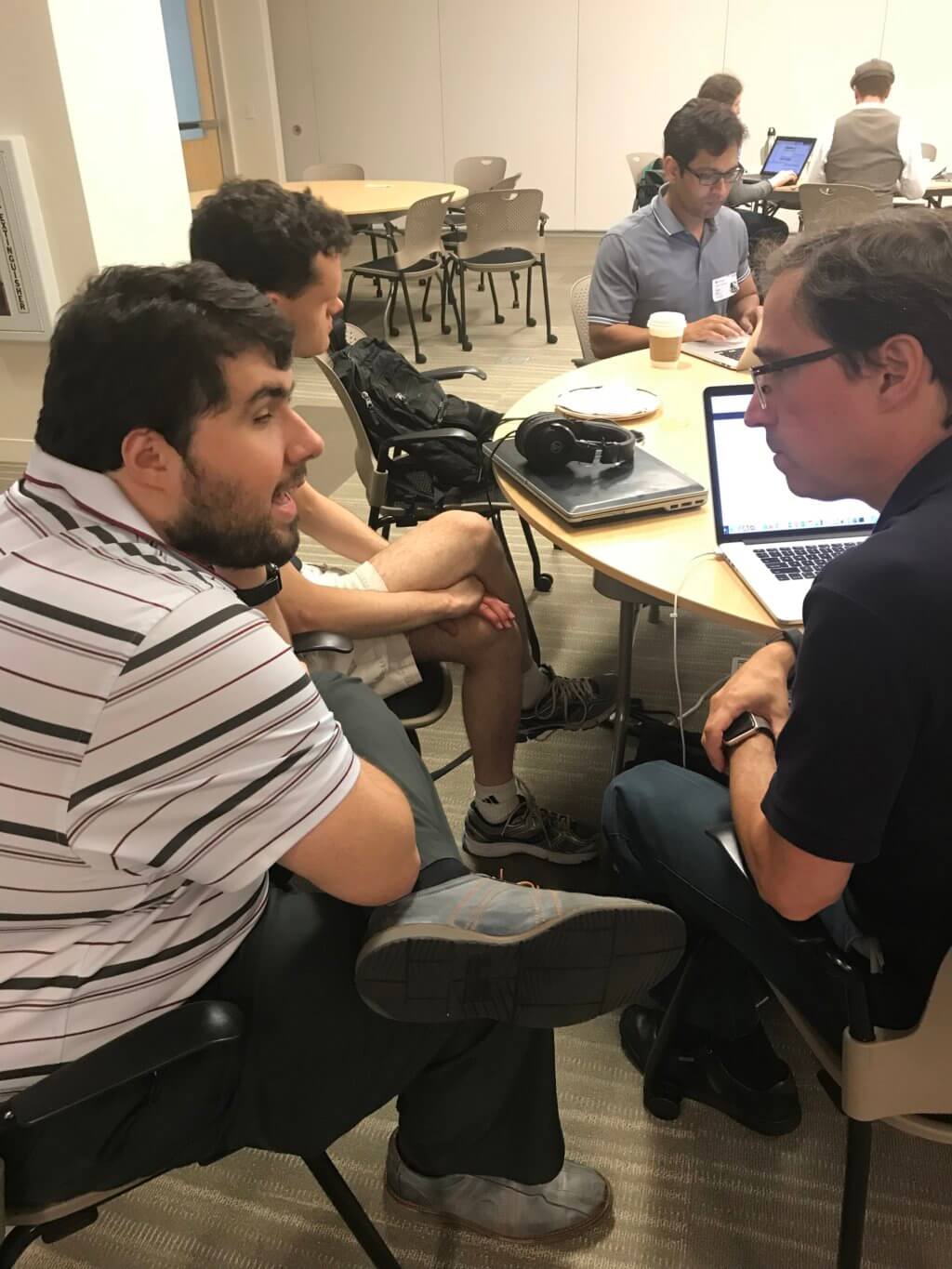 Sina leans into a conversation with a Benetech team member while sitting at a round table filled with laptops.