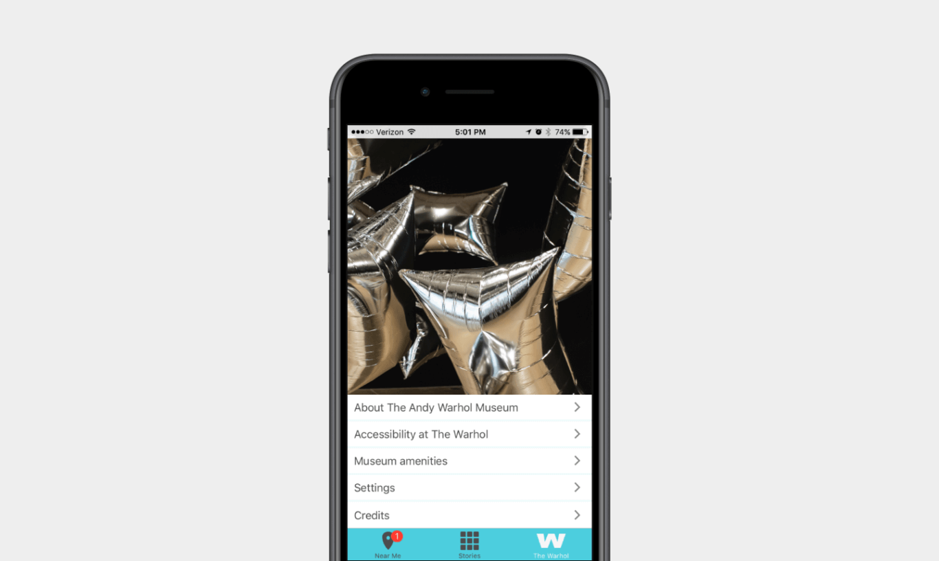 Half of a mobile phone screen features an image of reflective silver square balloons; the lower half lists 5 menu options: About the Andy Warhol Museum, Accessibility at the Warhol, Museum Amenities, Settings, and Credits.