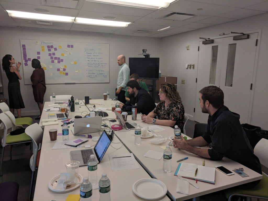 Sina and 2 other people sit around a group of 4 tables pushed together in meeting room. They look at white board where 3 other people stand reviewing post-its that cover the board. Laptops, notebooks, and coffee cups cover the surface of the tables.
