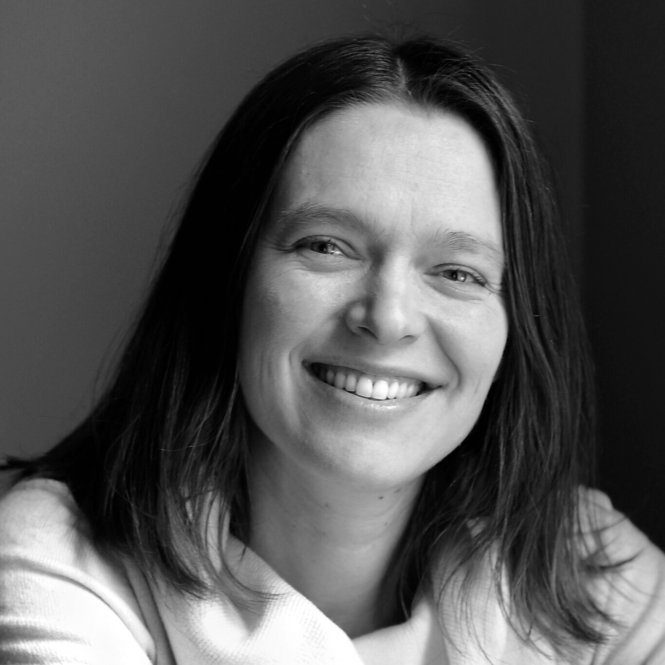 The headshot of a White woman with light skin, dark shoulder-length straight hair, and a wide smile. Her head tilted slightly to our right and her arms folded in front of her, resting on something out of the frame of the photo. She wears a white turtleneck sweater with a loose-fitting collar.