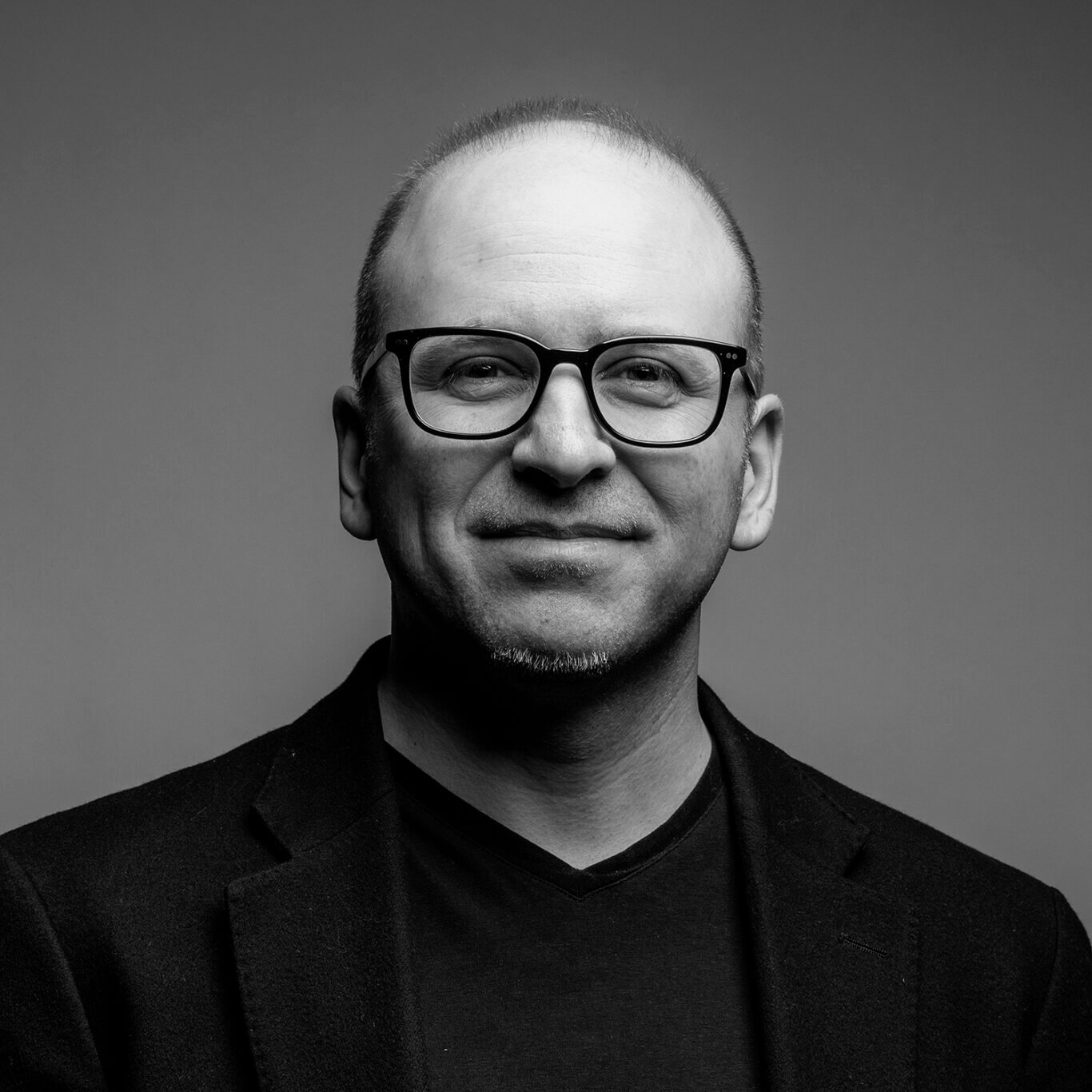 An up close headshot of a White man with light skin wearing black framed glasses. He smiles directly at us with a slight dimple on our left, and smile wrinkles around his eyes. He has thinning hair, subtle stubble, and a short white goatee. He wears a black v neck t-shirt under a black sports coat in front of a light grey background.