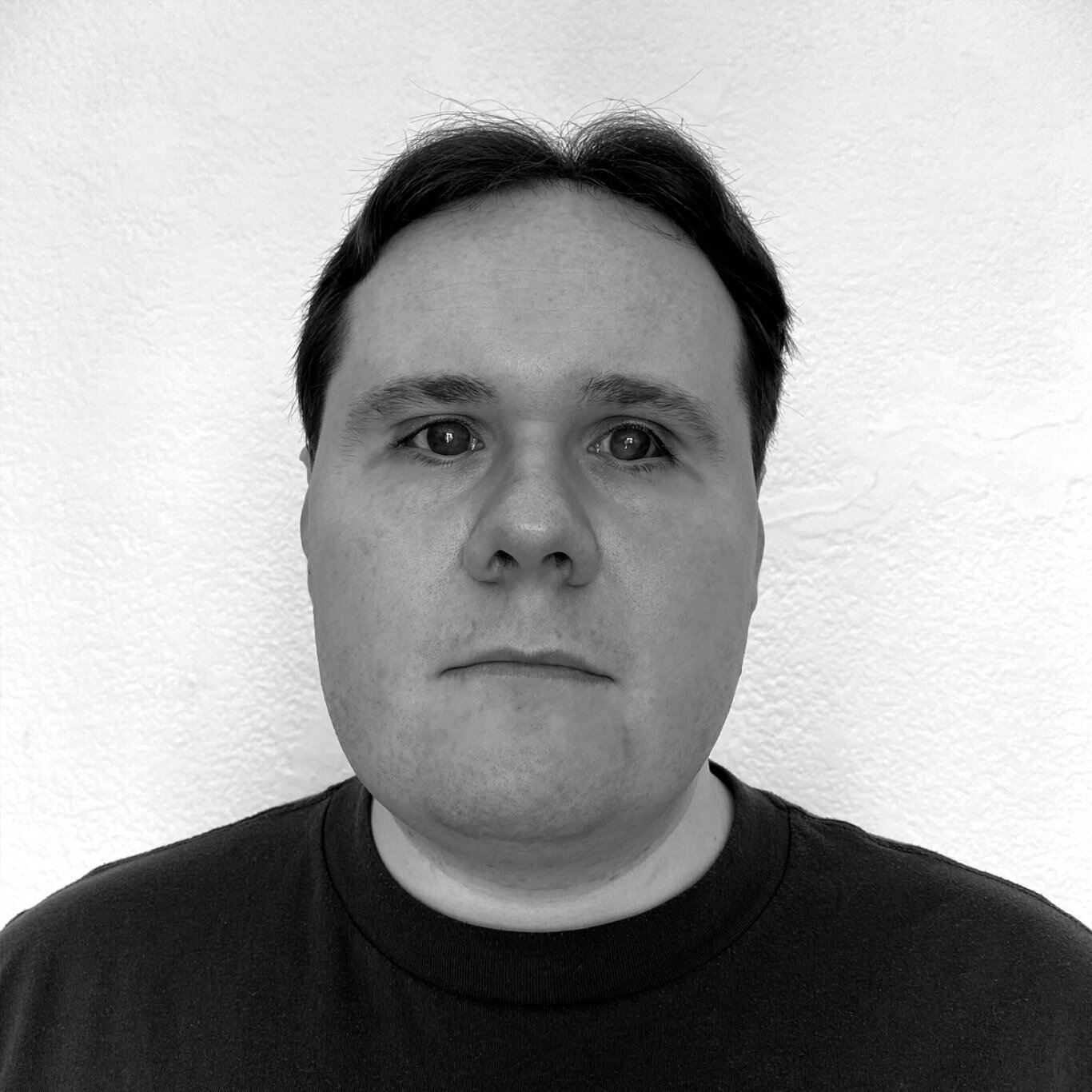 A closely cropped headshot of a White man with light skin and a neutral expression on his round face. His short dark hair is parted in the middle and slightly longer on top. He is wearing a dark t-shirt and his large eyes look past our right shoulder.