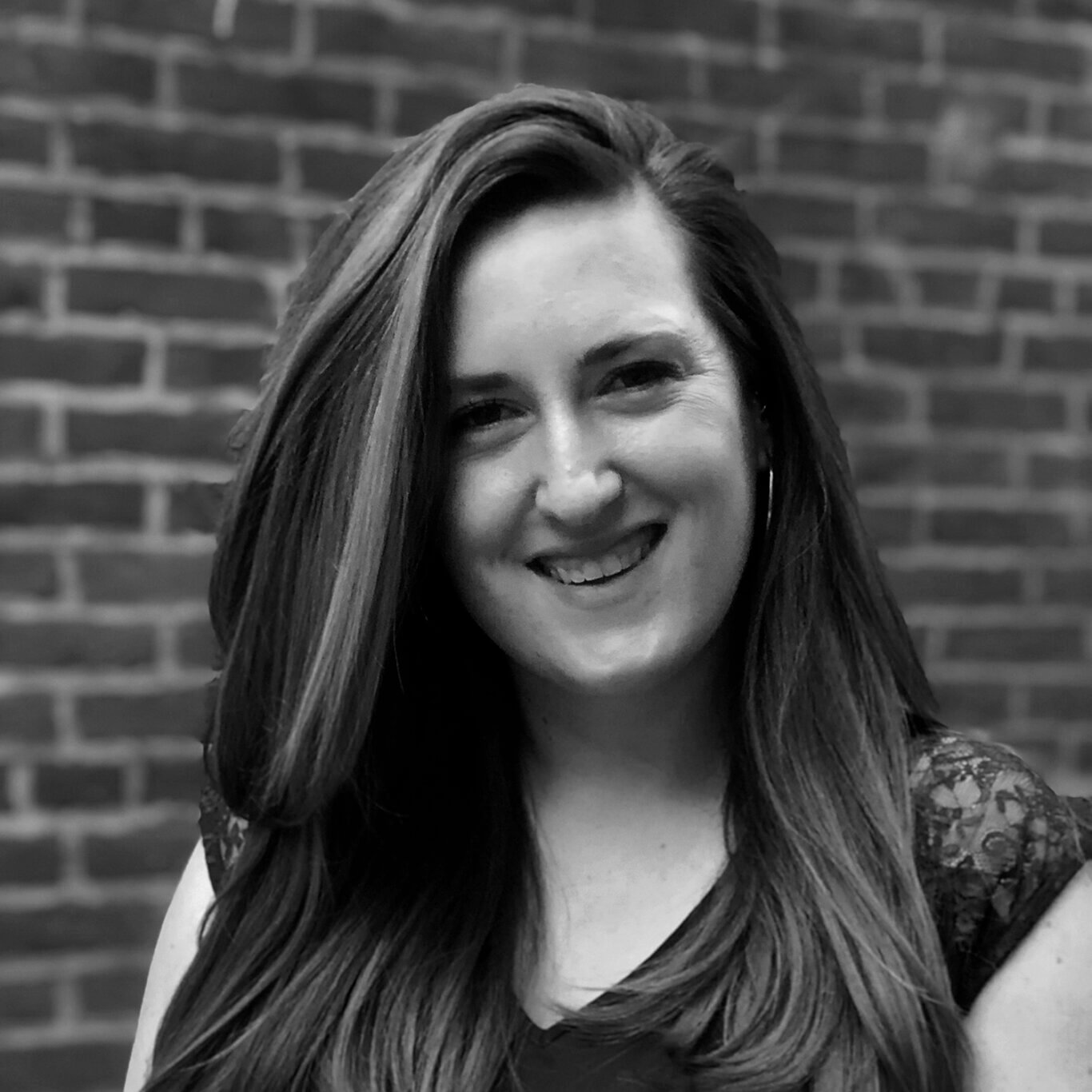 A black and white headshot of a White woman with light skin and voluminous medium tone hair shown from the chest up, her head tilted slightly to our left. She smiles widely, her eyes crinkling slightly. She wears a V neck shirt with lace capped sleeves, and thin silver hoop earrings. Behind her is a brick wall.