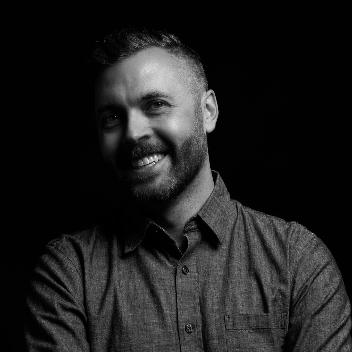 A black and white headshot of an Indigenous Métis man with light skin and a toothy grin. Pictured from the chest up, head tilted slightly to our left, and smile wrinkles surround his eyes. He has short, quaffed salt and pepper hair and full beard. He wears a light grey button-down shirt in front of a dark background.