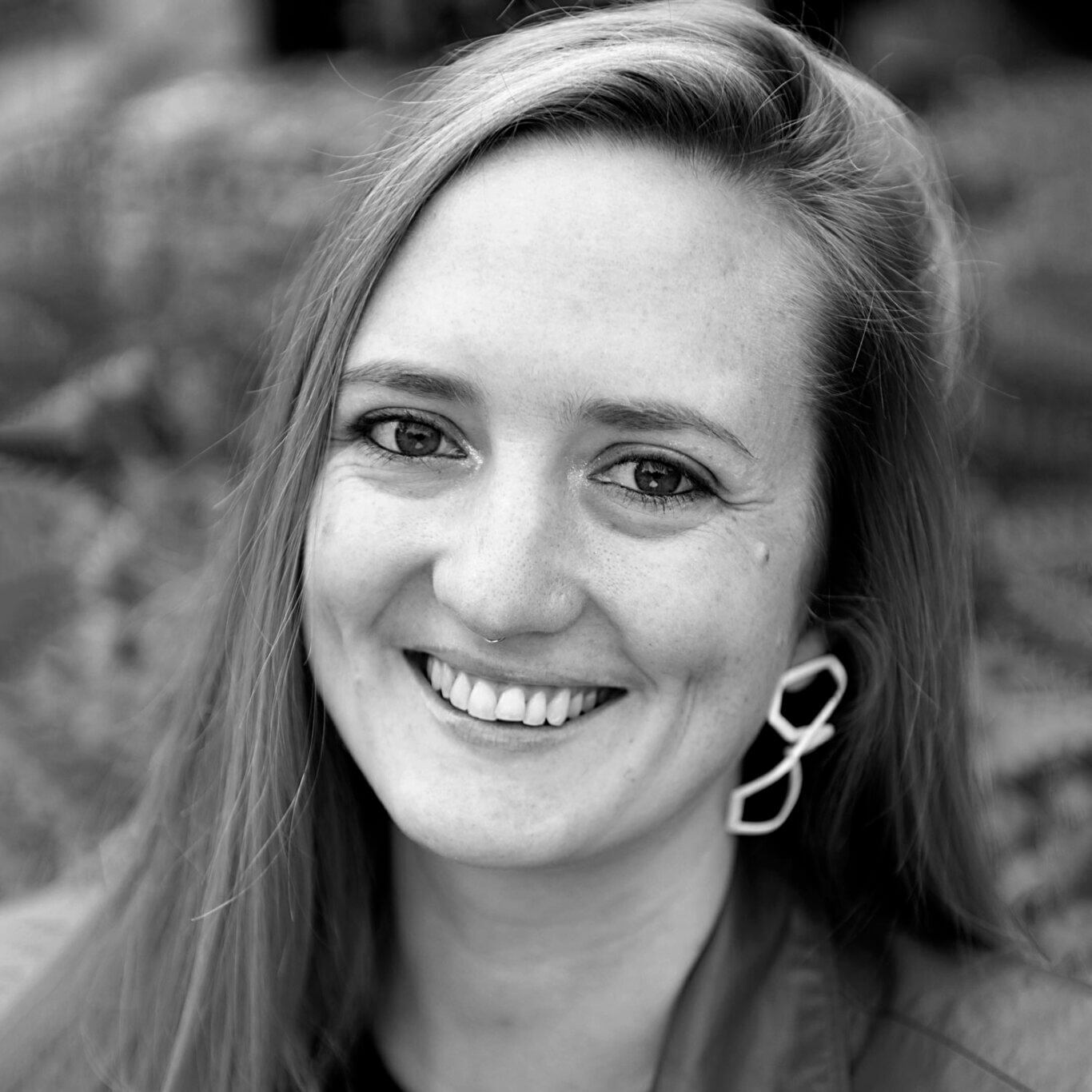 A closely cropped black and white headshot shows a White nonbinary femme with light skin tone and two large dimples smiling widely at us. At the edge of their smiling, crinkling eyes is a hint of glitter. Their long straight hair is tucked behind their left ear, on our right showing a large white geometric earring.