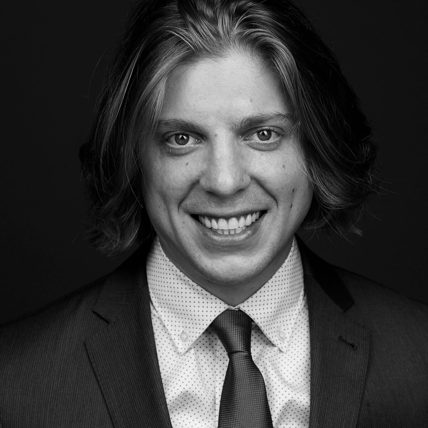 A closely cropped black and white headshot of a White man with light skin and a wide smile. His medium toned chin length hair is swooped back. He wears a sharp gray suit, a pressed white shirt with tiny dots, and a dark tie. He has a few small moles scattered primarily on our right, his left, cheek.