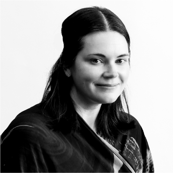 A black and white headshot of a White woman in her late thirties. She has light skin, shoulder-length straight dark hair that is half pulled back and parted in the middle. Viewed from the chest up, she is wearing a dark wrap dress with a painterly pattern, and sits in front of a white background.